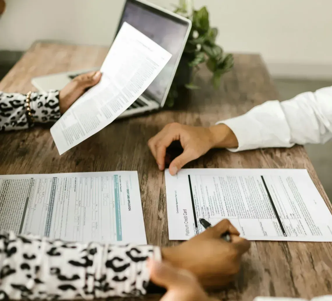Two people at a wooden desk reviewing documents, one holding a paper while the other holds a pen near a laptop.