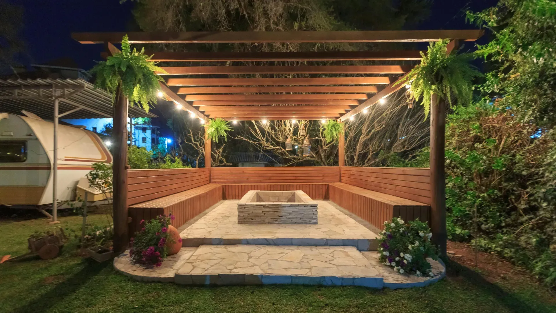 Wooden pergola with bench seating surrounding a fire pit, lit by string lights at night.