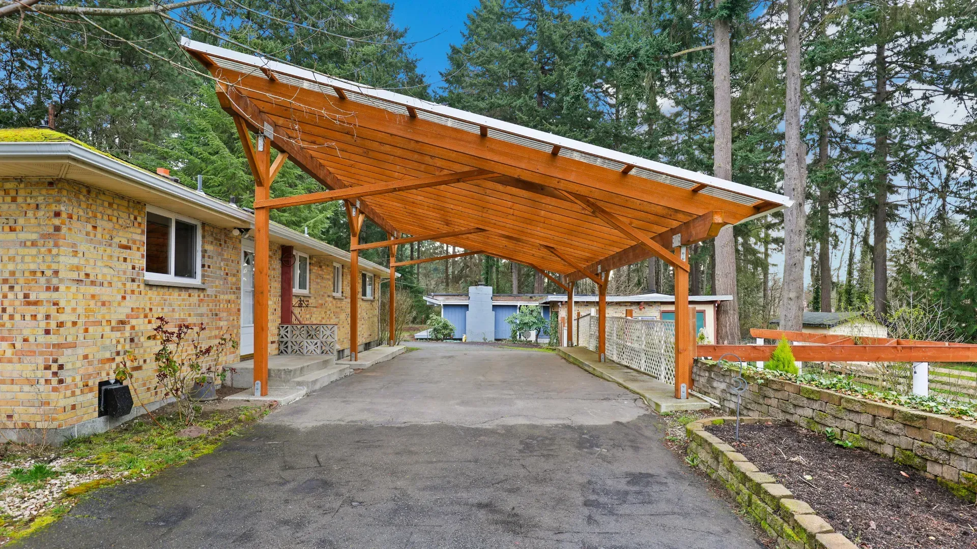 Carport with wooden frame over a driveway, next to a brick house and trees.