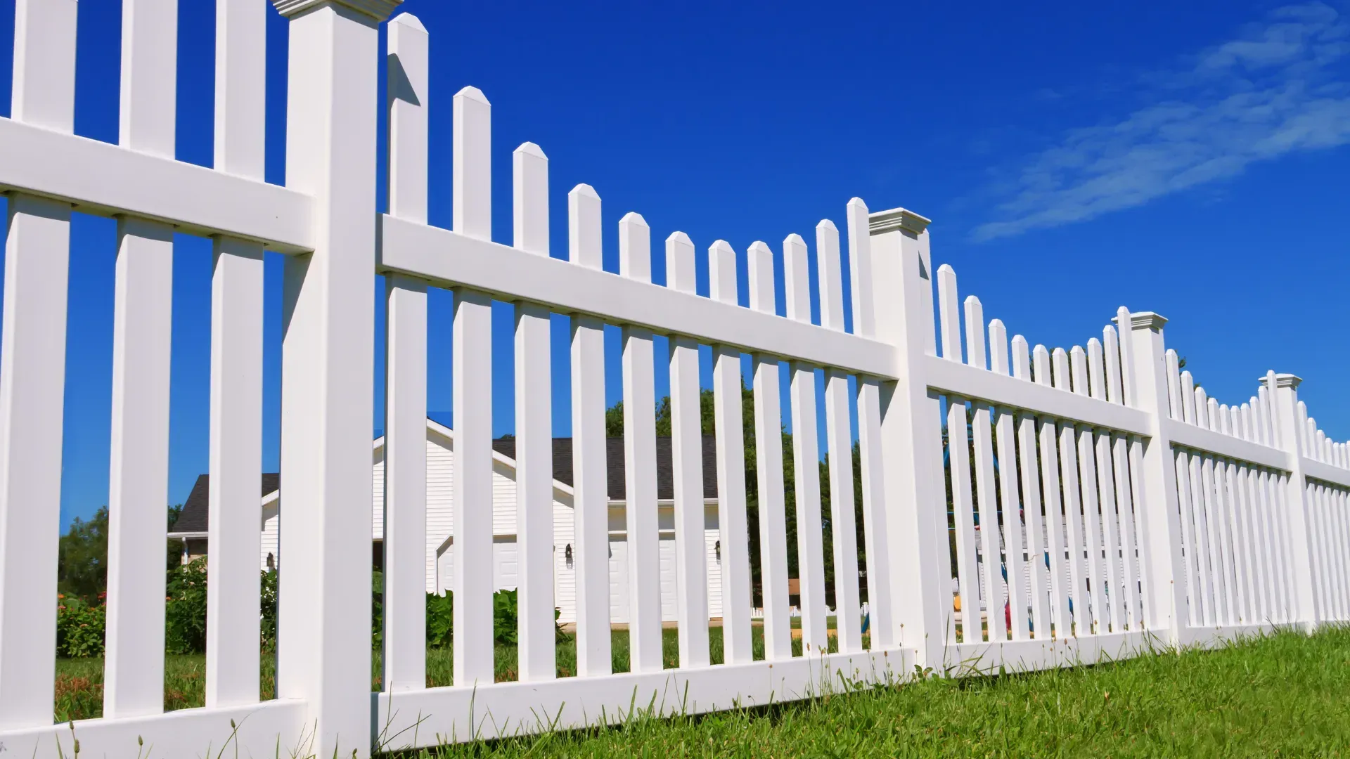 White picket fence against a bright blue sky, with a house visible in the background.