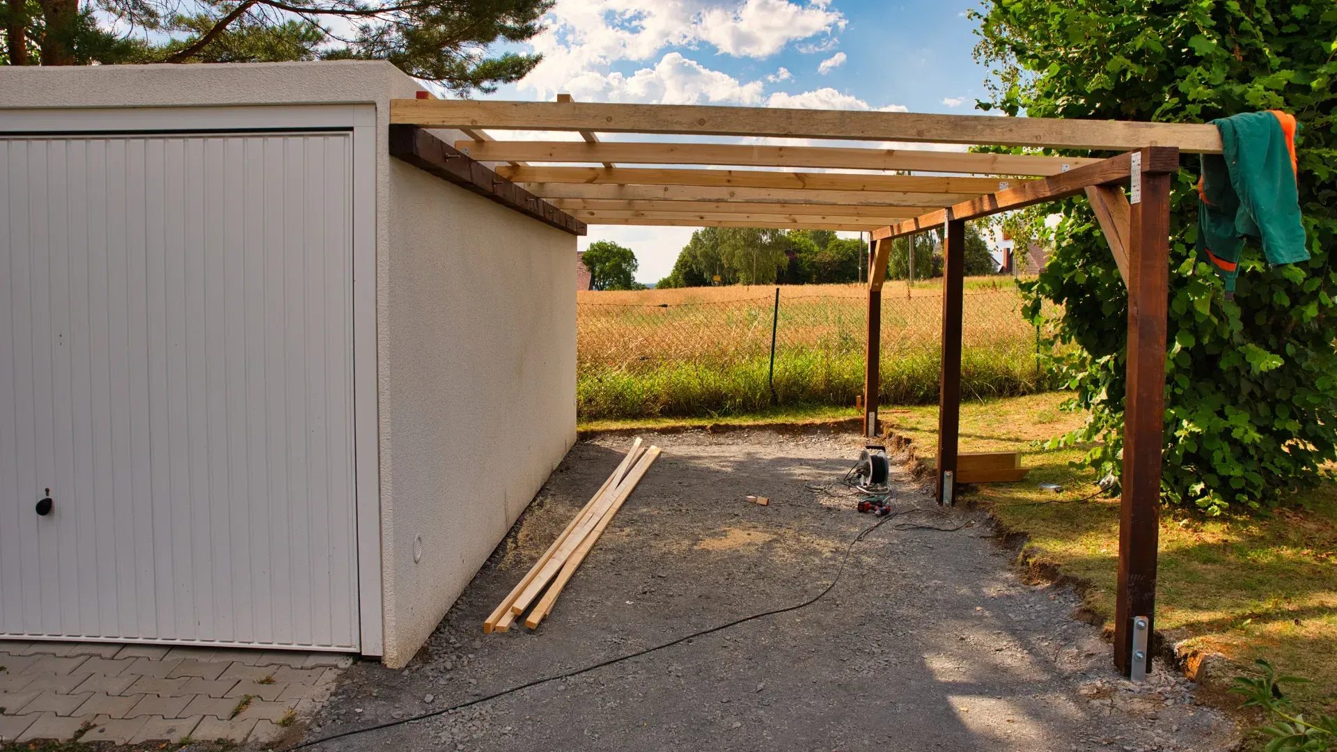 Carport attached to garage with wooden beams and posts on gravel.