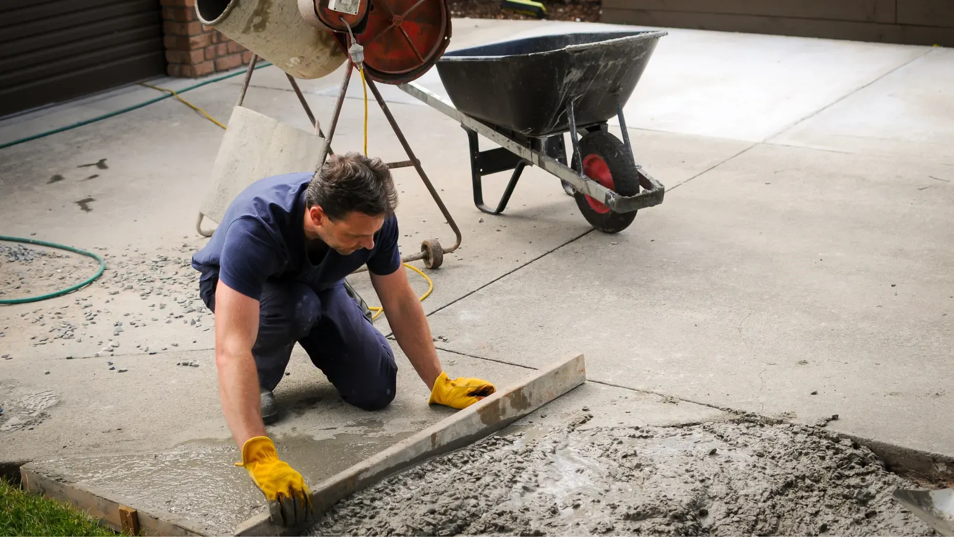Man leveling fresh concrete with a level; yellow gloves; cement mixer and wheelbarrow in background.