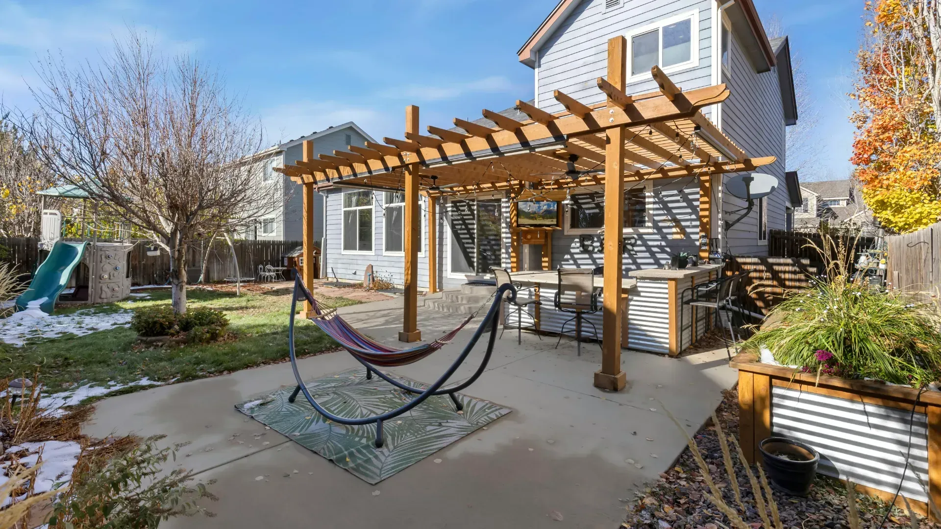 Backyard with pergola, hammock, and a two-story house with gray siding. Sunny day.