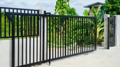 Black metal gate with vertical bars, concrete pillars, and lush green foliage.