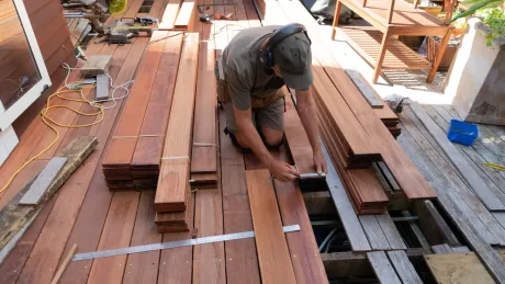 Man kneeling, measuring wood planks on a partially built deck. Wearing safety gear, outdoors.