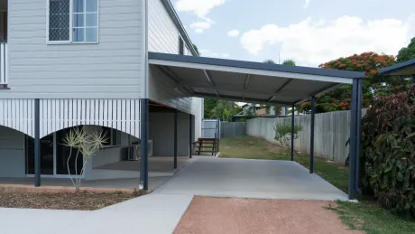 Carport attached to a light grey house, with a concrete driveway and small yard.