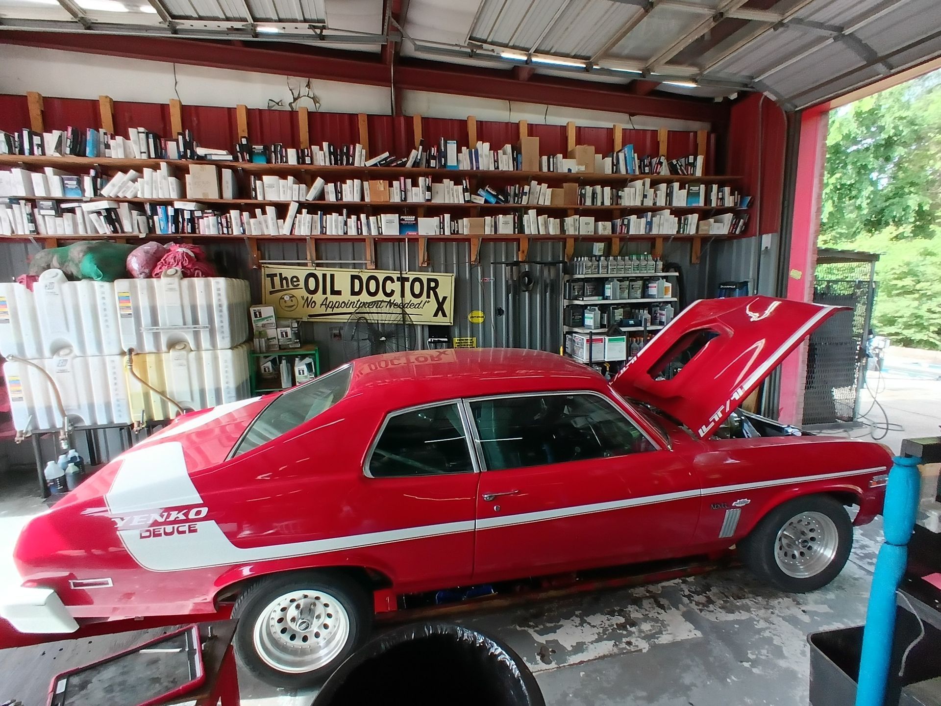 Red classic car in a garage, hood open. White racing stripe, shelves of supplies in background.