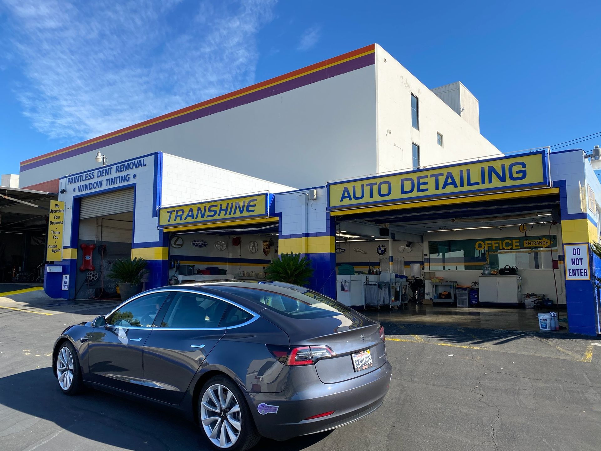 A gray tesla model 3 is parked in front of an auto detailing shop.