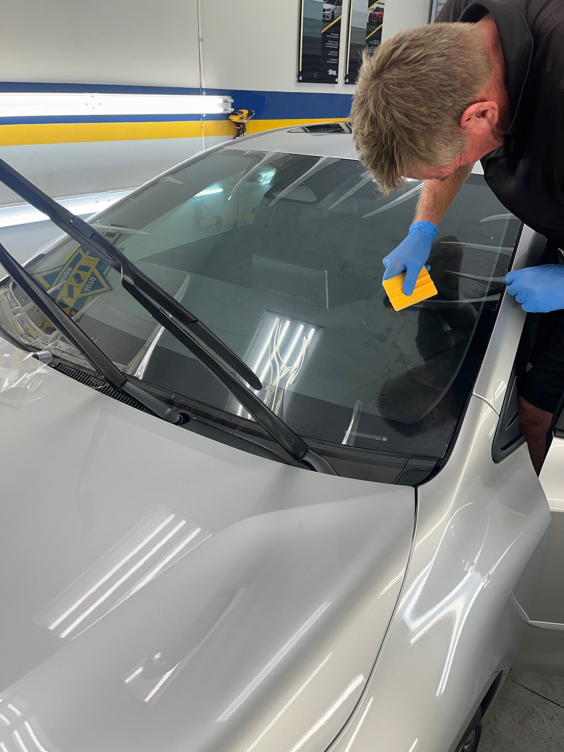 Man applying tinted film to a car windshield in a shop using a squeegee, wearing gloves.