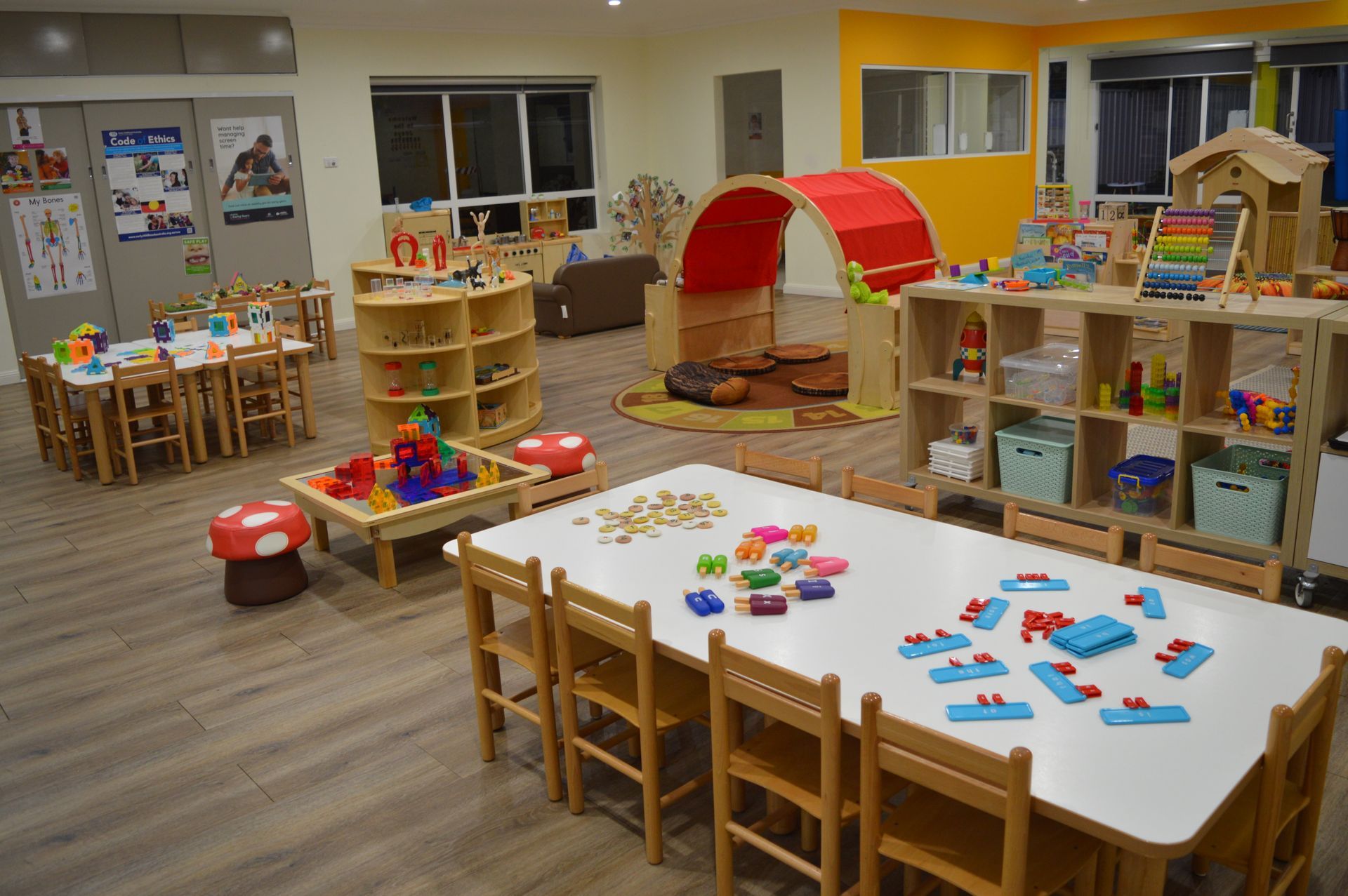 A bright, colorful preschool classroom filled with wooden tables, chairs, toys, and a red arched play structure.
