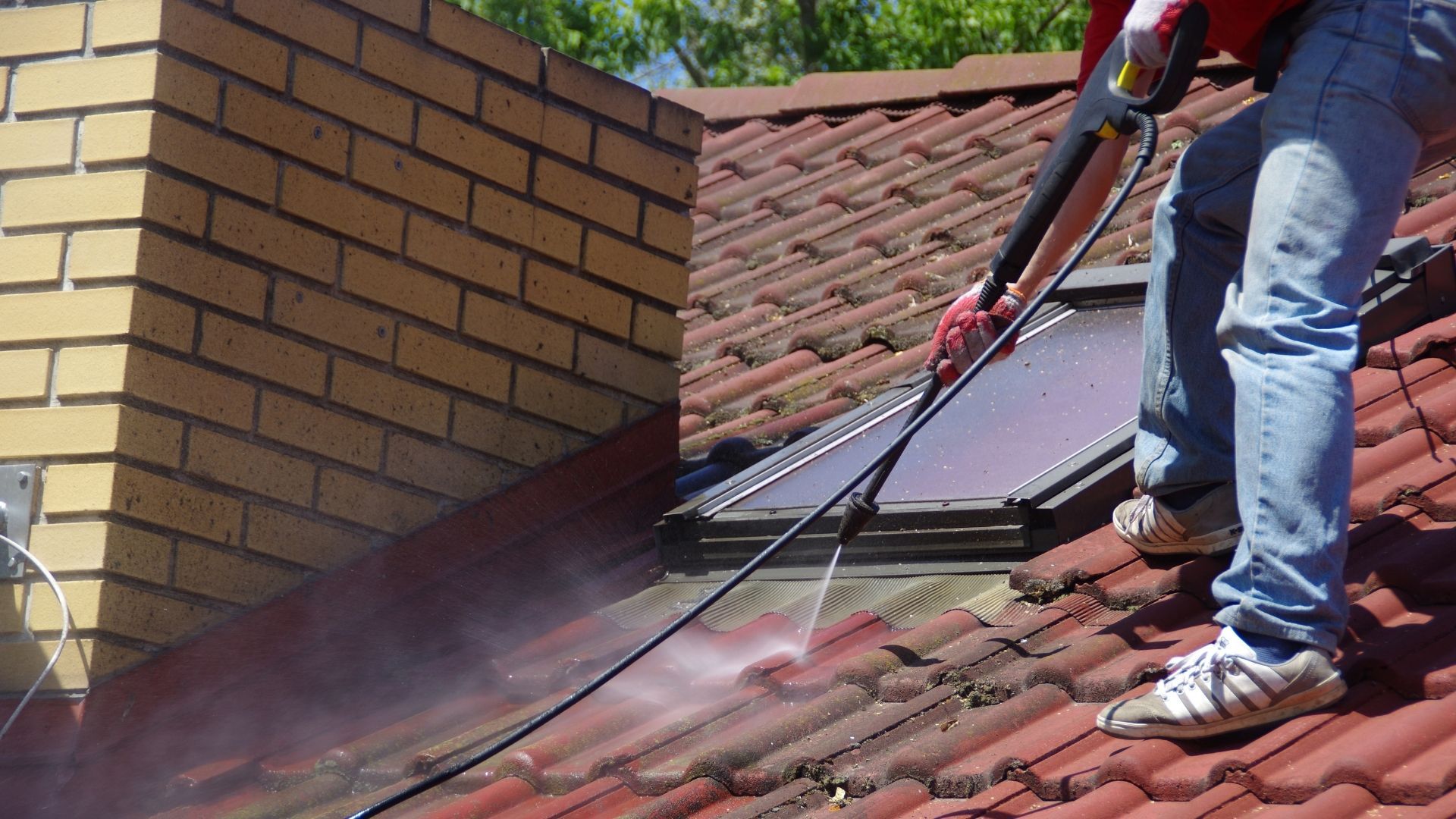 Person pressure washing a red tile roof, near a brick chimney and skylight.
