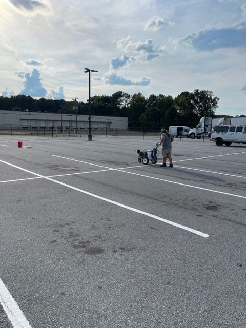 Person painting parking lines in an empty parking lot with a striping machine. The lines are white; the sky is cloudy.