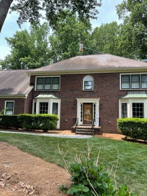 Brick two-story house with a brown roof and green bushes lining the lawn. The door has an arched top and is flanked by lights.
