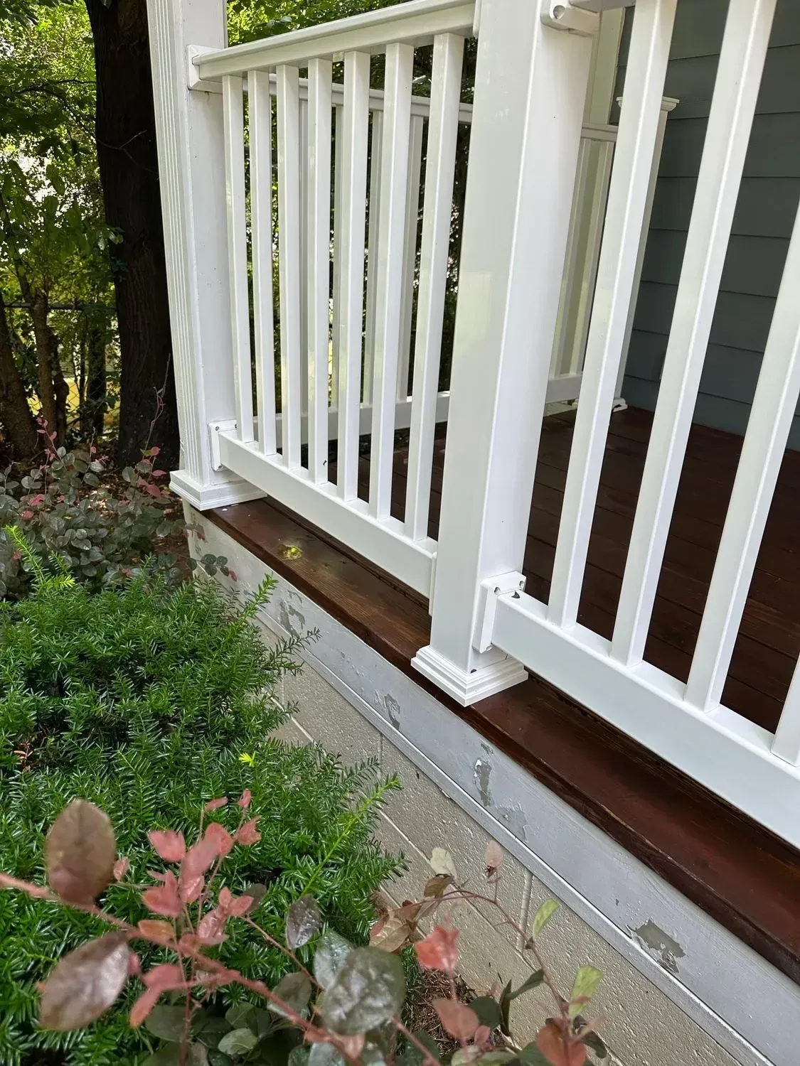 White porch railing with a dark brown wooden base next to shrubs.
