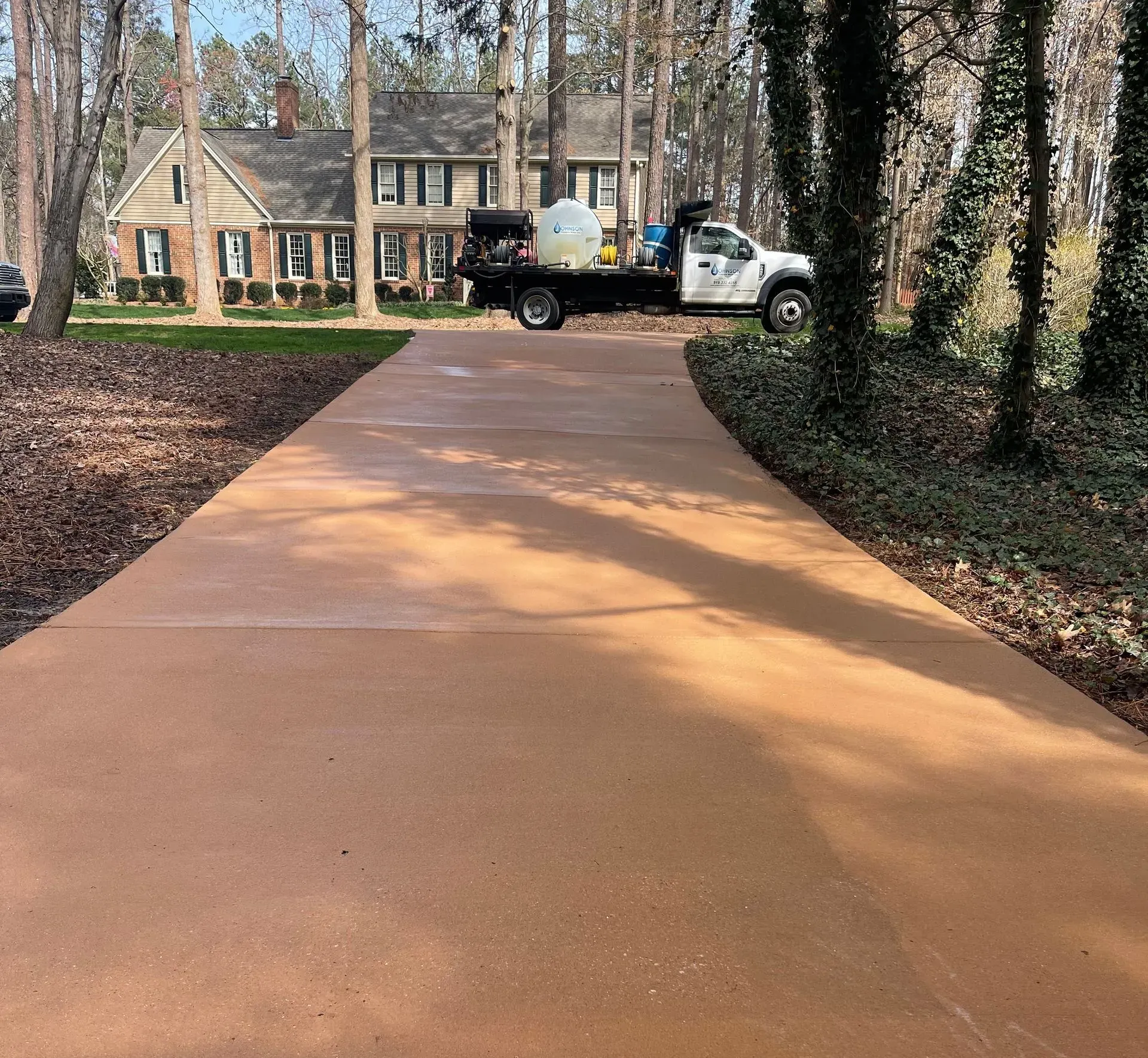 Brown concrete driveway with a spray truck in front of a two-story brick house surrounded by trees.