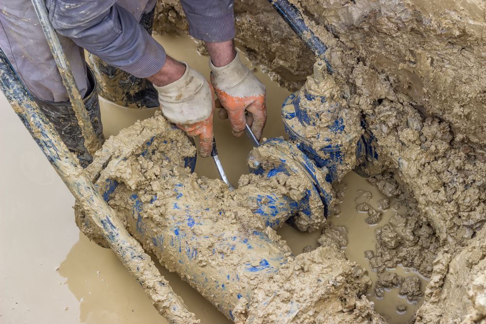 A Man is Working on a Muddy Pipe in the Dirt — Hoges Plumbing In West Wollongong, NSW