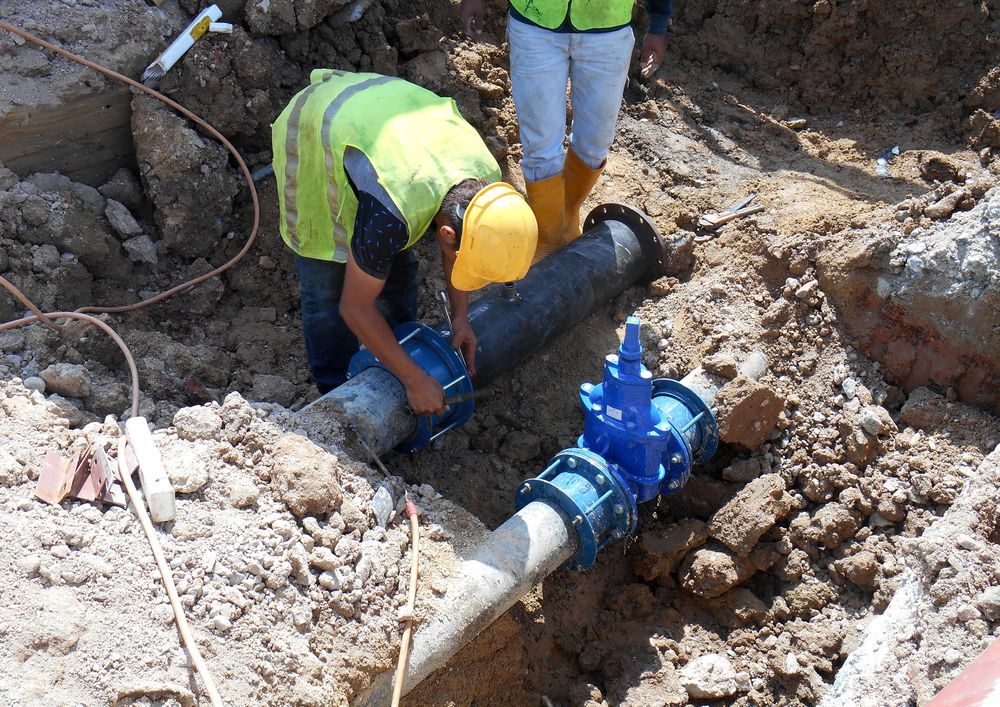 A Man in a Yellow Hard Hat is Working on a Pipe in the Dirt — Hoges Plumbing In Kiama, NSW