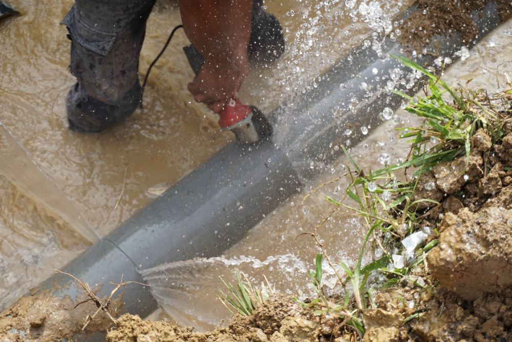 A Man is Cutting a Pipe With a Grinder in the Mud — Hoges Plumbing In West Wollongong, NSW