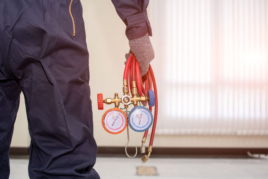 Person holding red HVAC gauges with hoses, standing indoors near a window.