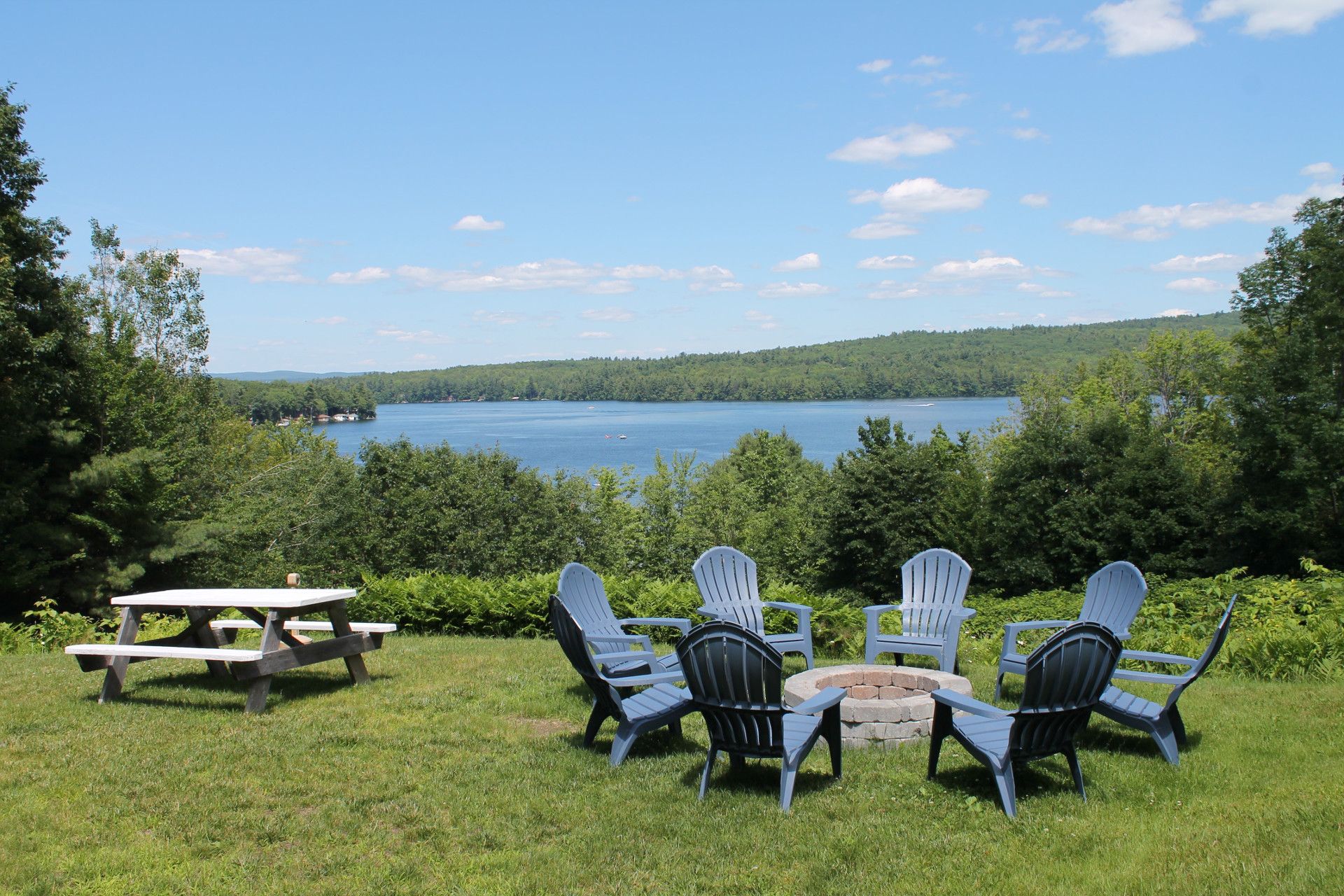 A lakeside view with a picnic table and Adirondack chairs circling a fire pit on a grassy hill. Blue sky.