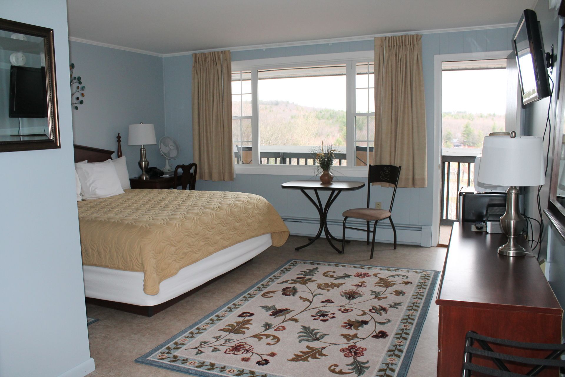Bedroom with a bed, desk, small table, and access to a balcony. Beige and blue color scheme, mountain view visible through the windows.
