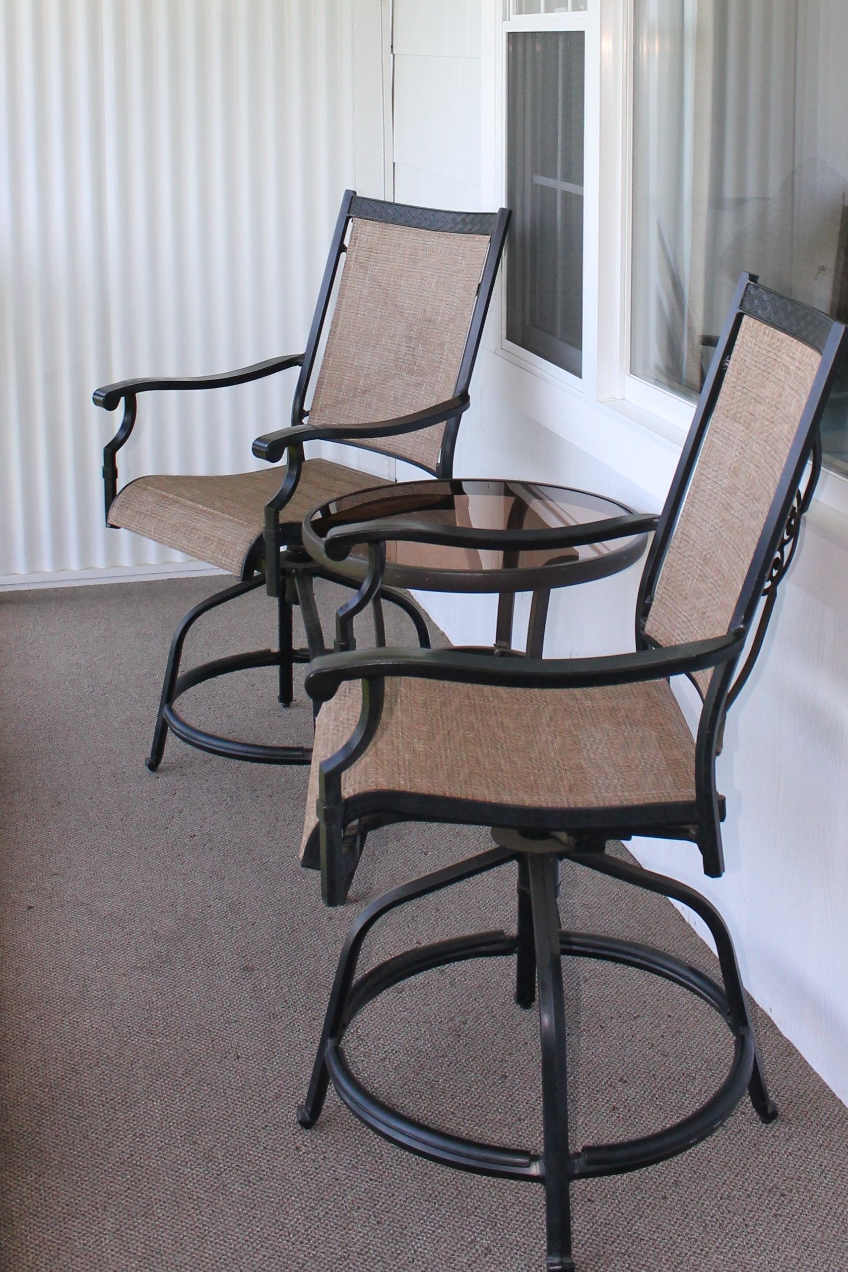Two metal chairs with woven seats and a small table on a porch, set against a window.