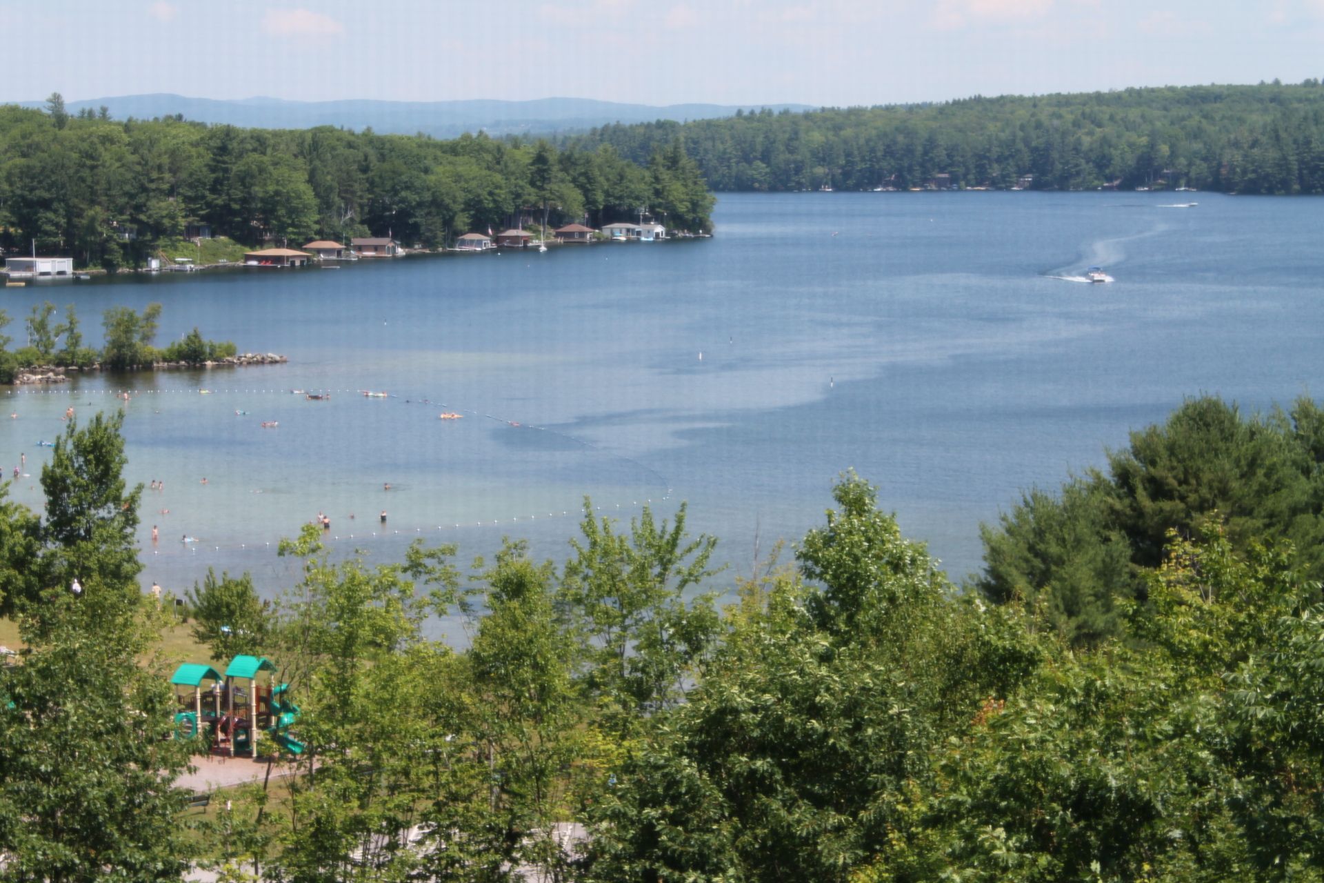 A scenic lake view with houses along the shore, trees surrounding the water, and a playground in the foreground.