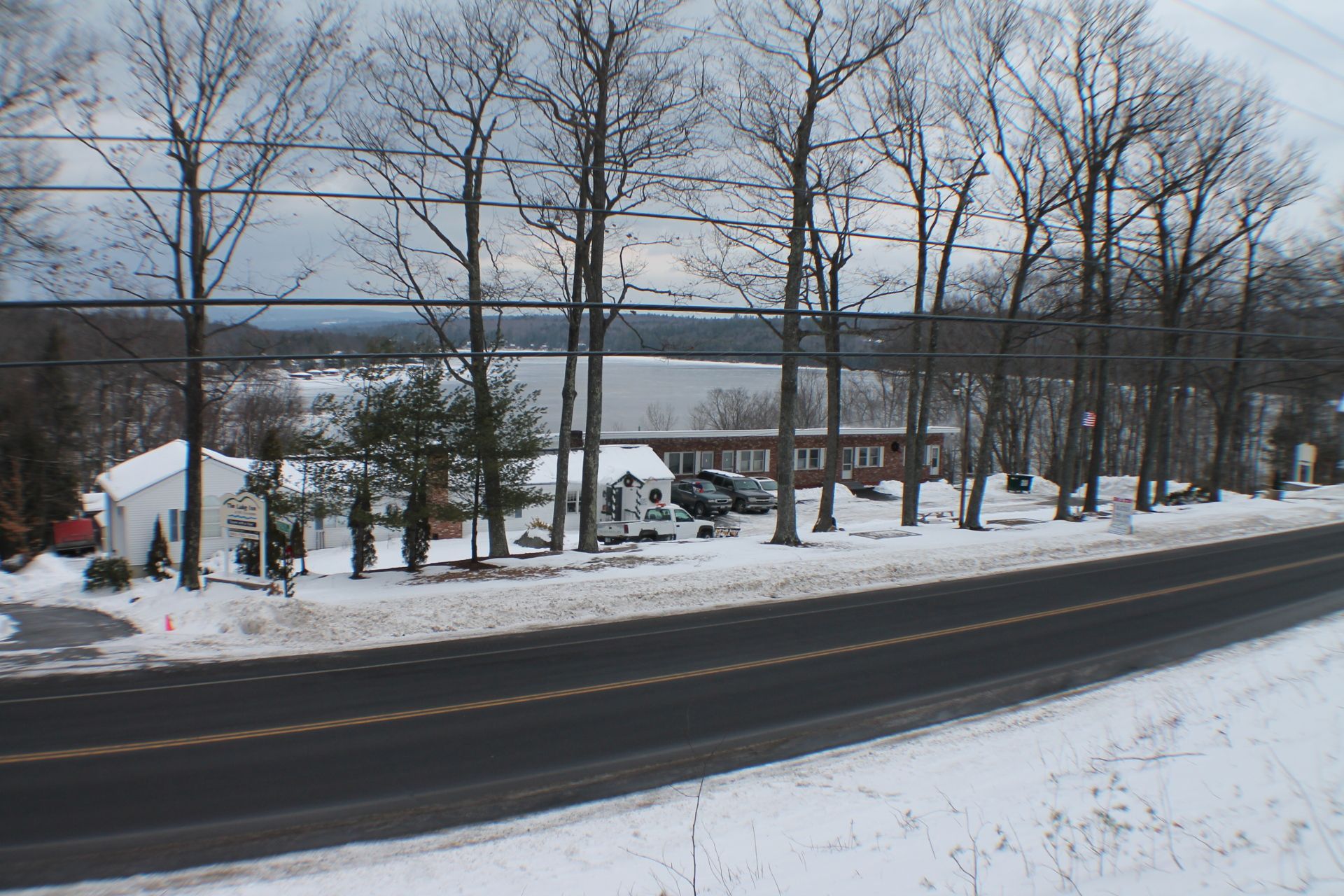 Snowy roadside view with a motel and bare trees; a lake is visible in the distance under a cloudy sky.
