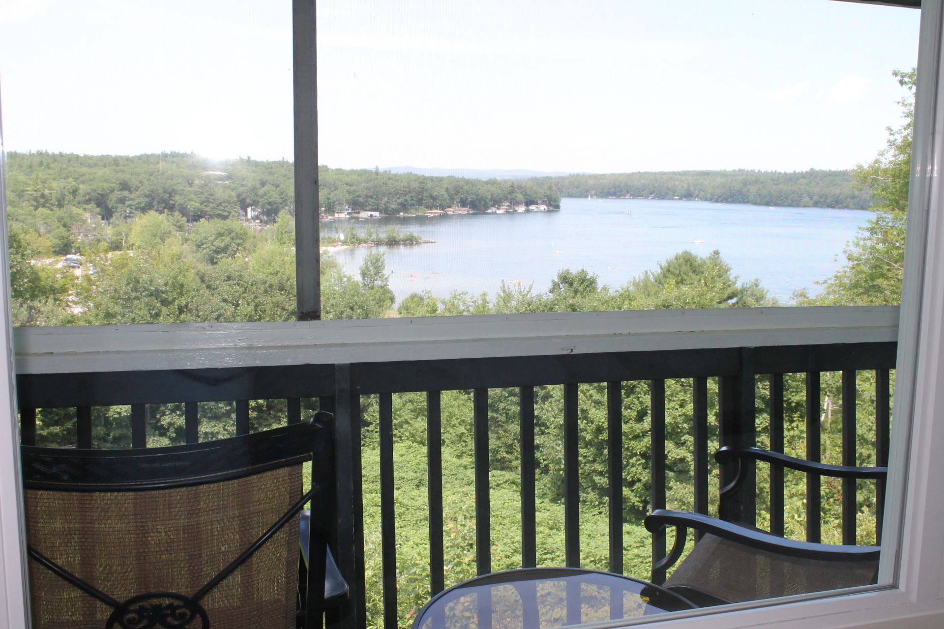 Balcony overlooking a lake. Dark railing and chair in foreground; trees and water in view beyond the railing.