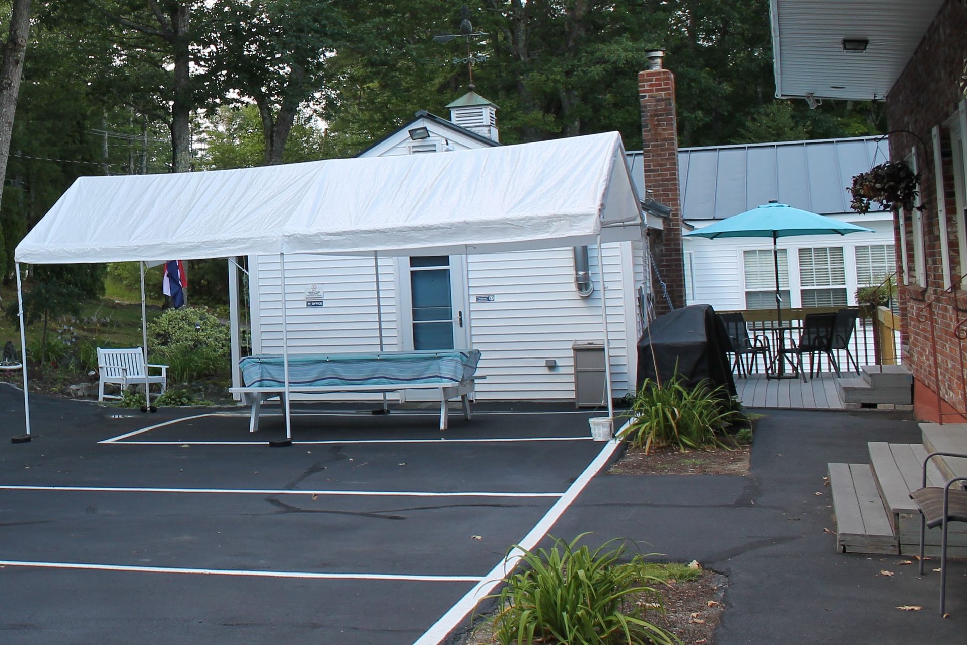 A small white building with a white canopy and parking in front. Another building with an outdoor table and umbrella is visible behind it.