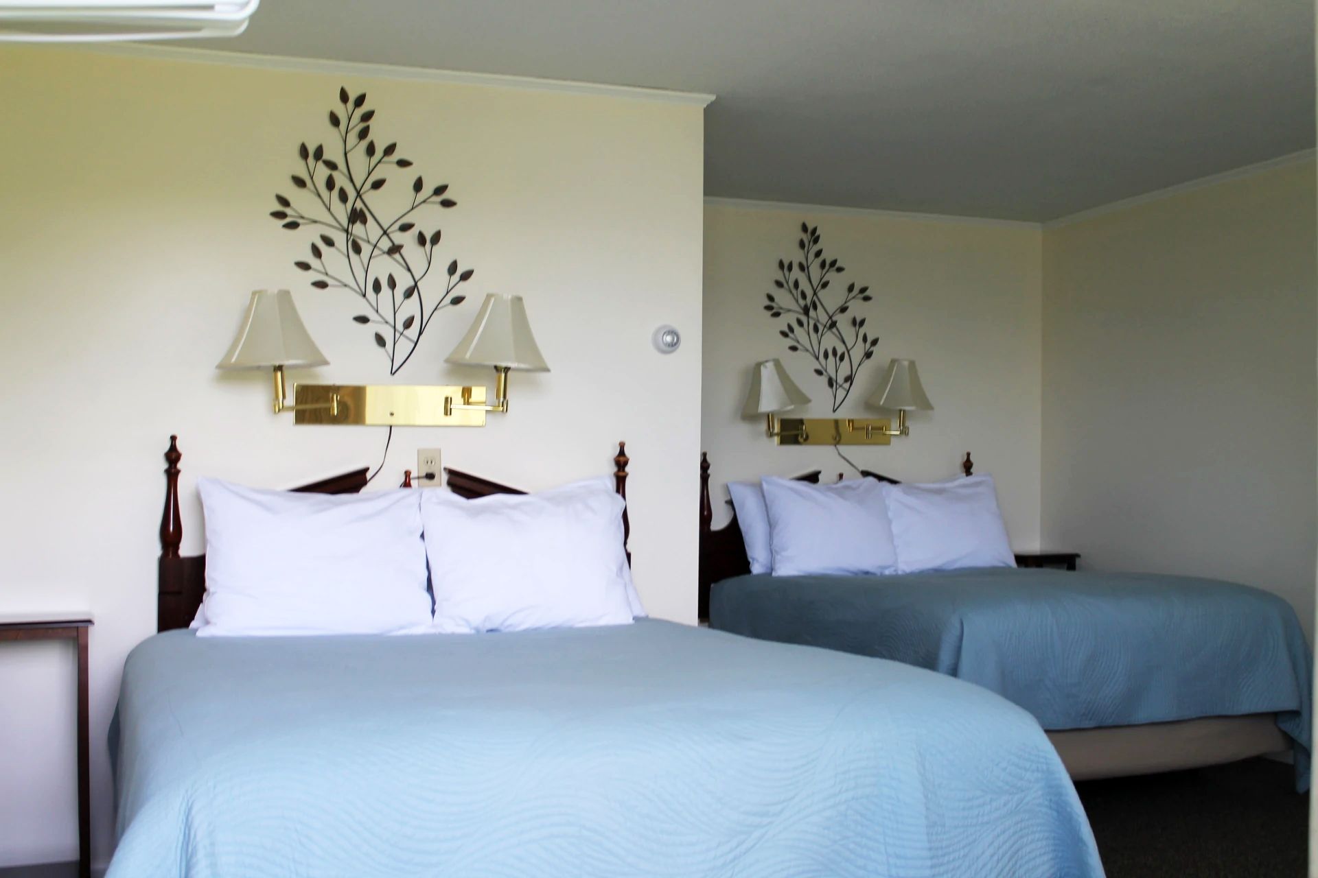 Classic Double Room with two motel beds with blue bedding, framed by wood headboards and wall sconces, against beige walls with decorative black leaf decals.