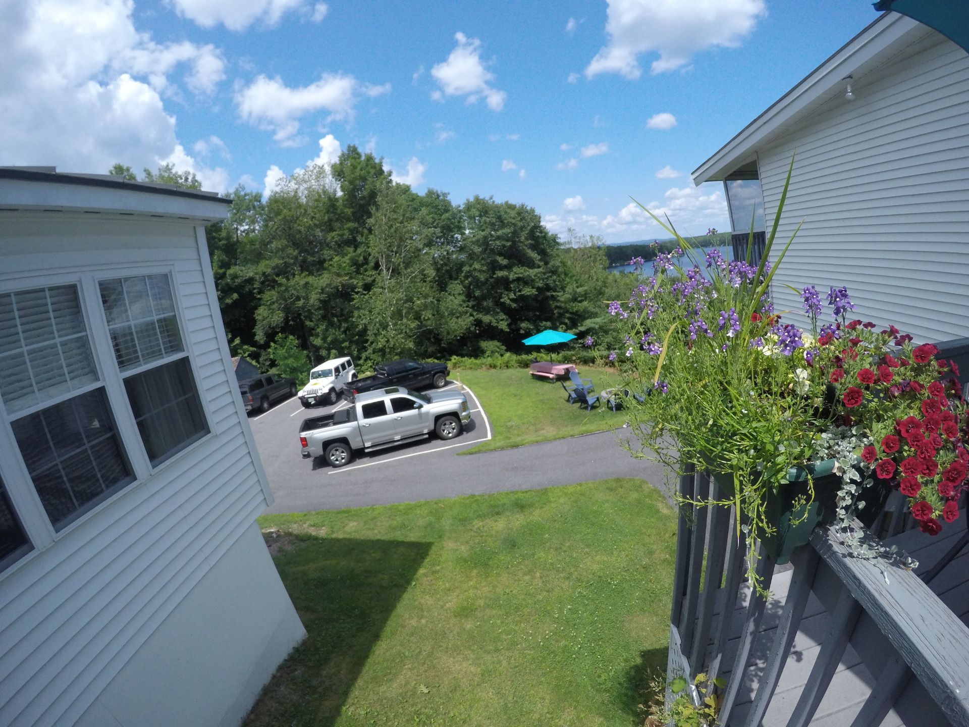 View from a balcony of a courtyard with parked cars, trees, and flower boxes, under a bright blue sky with clouds.