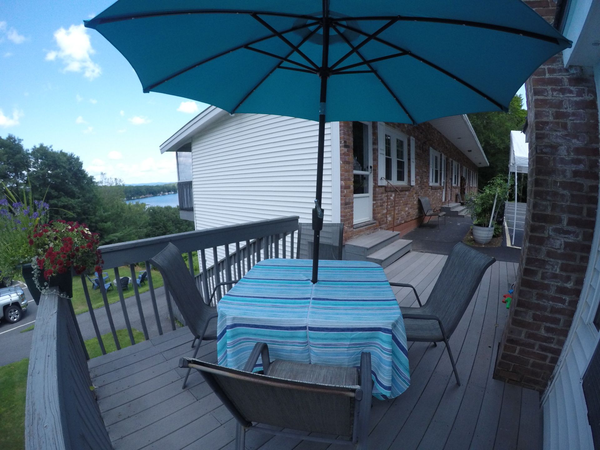 A deck with a table and chairs, covered by a blue umbrella. Houses and trees are in the background.
