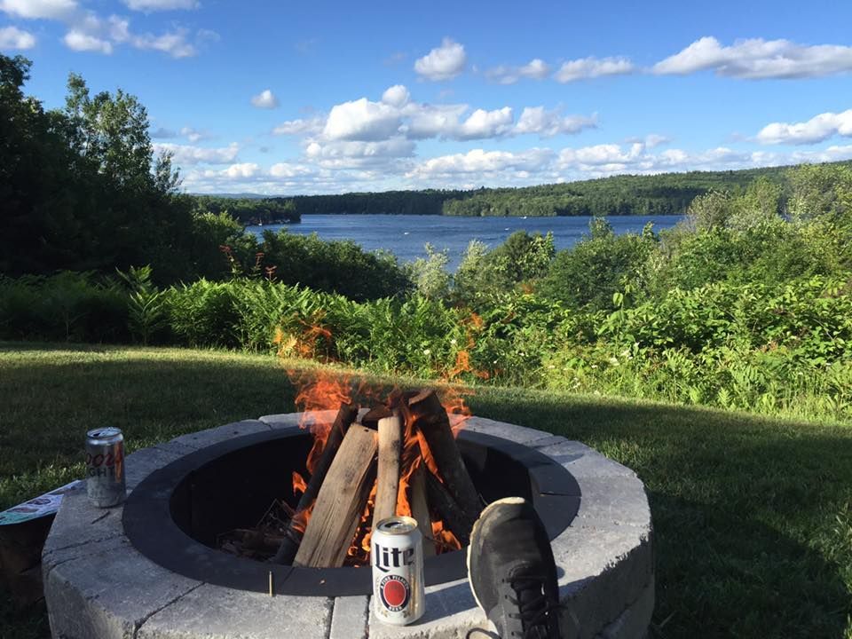 A campfire burns in a stone fire pit on a grassy lawn overlooking a lake on a sunny day. Beer cans sit nearby.
