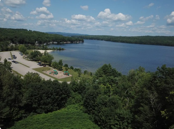 Aerial view of a lake with a beach, parking lot, and playground. Green trees surround the shore under a blue sky with clouds.