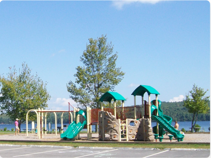 Playground by a lake under a blue sky.  Features swings, slides, climbing structures, and a tree.  A person stands near the swings.