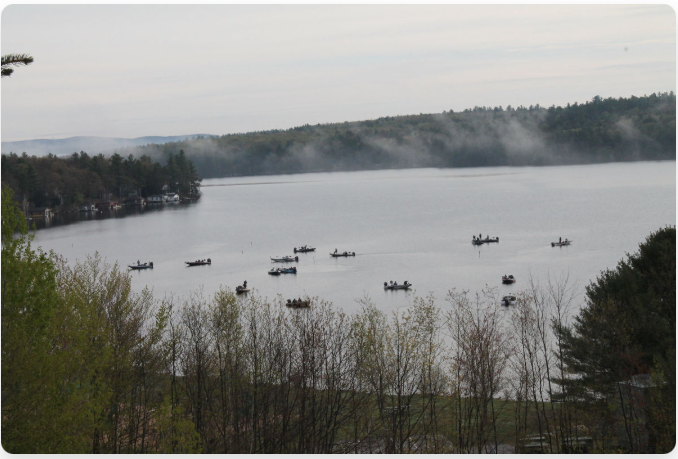 Boats scattered across a calm Lake Sunapee, a misty morning scene. Trees line the shores under an overcast sky.