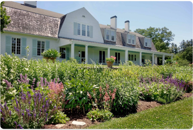 The Fells large white house with a long porch, light blue shutters, and a vibrant garden in front.