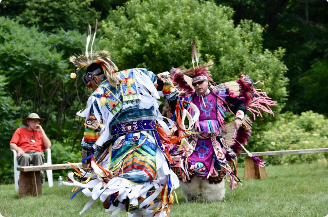 Two Native American dancers in colorful regalia perform outdoors. A person watches from a chair in the background.