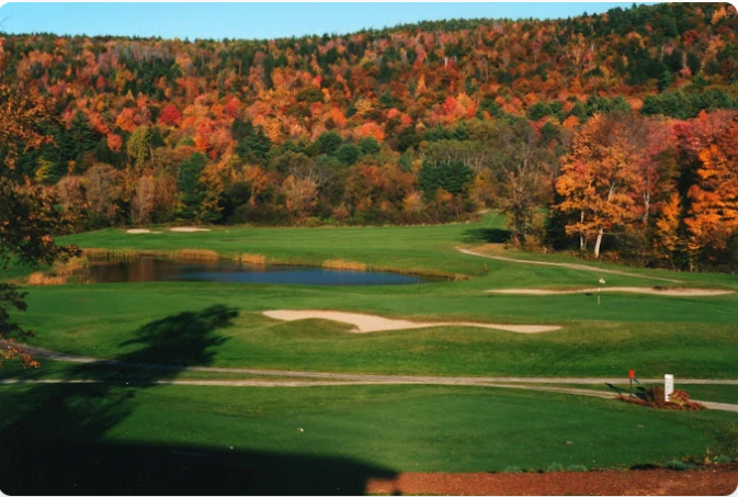 Golf course with a green fairway, sand traps, and a pond, set against a backdrop of colorful autumn trees.