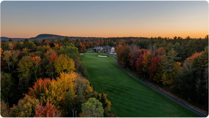 Golf course fairway lined with colorful autumn trees at sunset. A house is visible in the distance.
