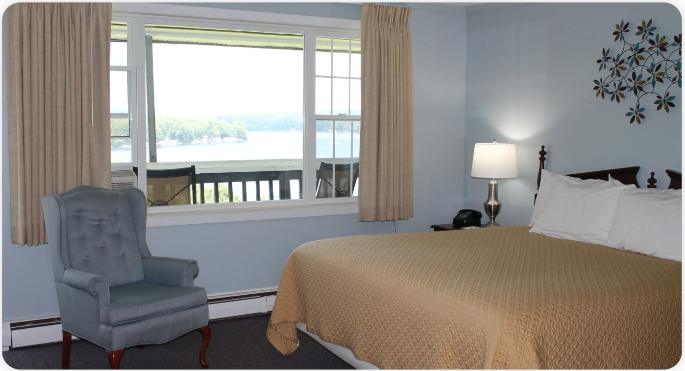 A hotel room with a bed, a window overlooking a lake, and a blue armchair. Soft beige curtains and a decorative wall piece are also visible.