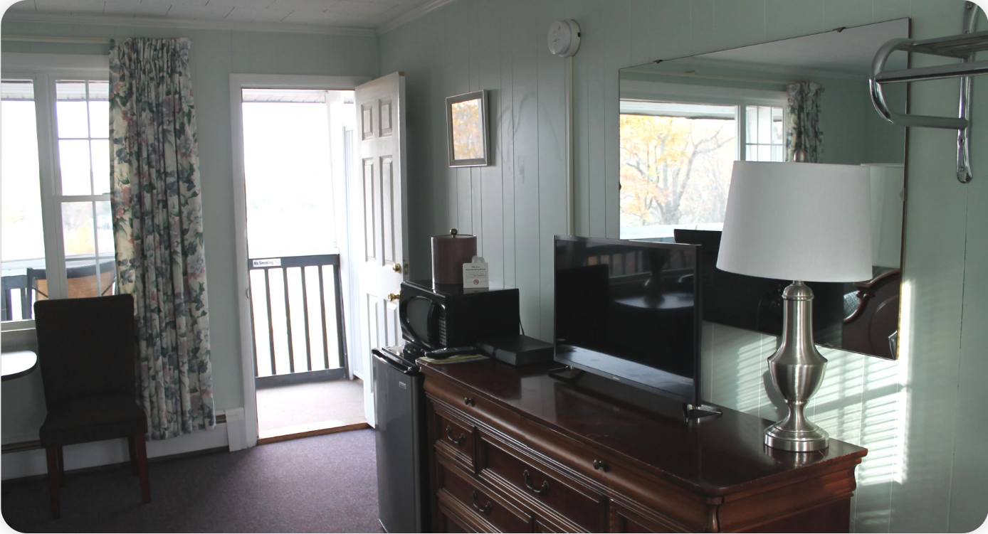 Premium Queen Anne Balcony hotel room interior with a dresser, TV, large mirror, open door to a balcony, and windows with patterned curtains.