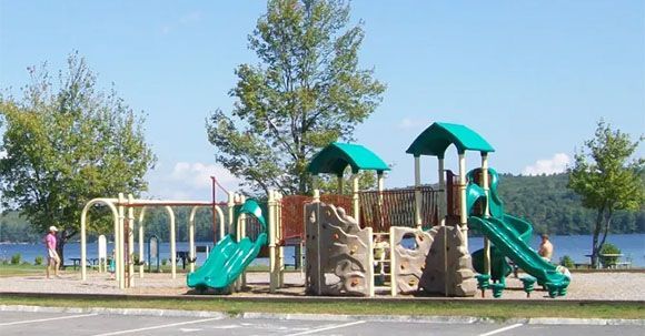 Playground with green slides, swing set, and climbing structure next to a lake.