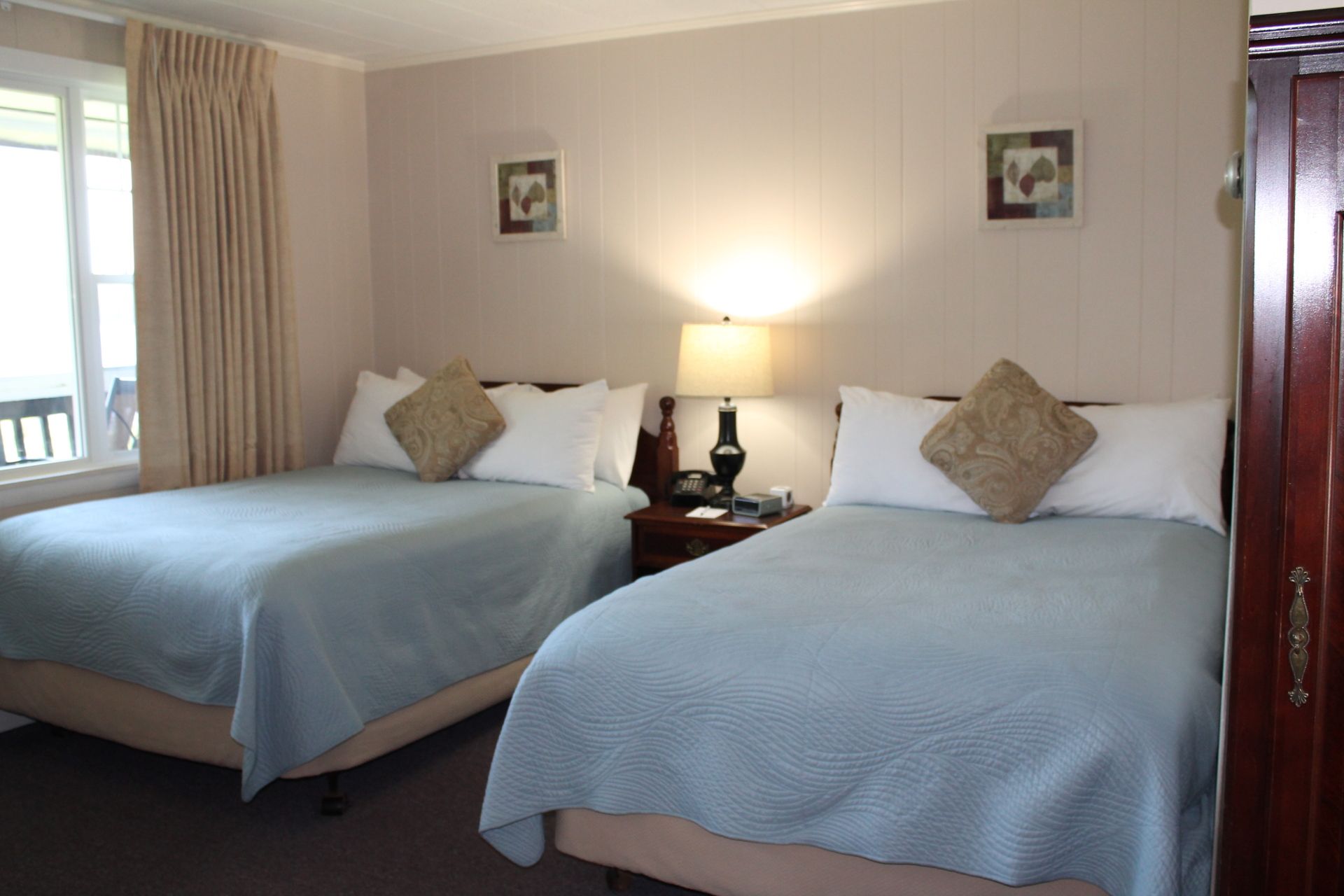 A hotel room with two beds covered in light blue quilts. A lamp sits on a nightstand between them. Beige walls.