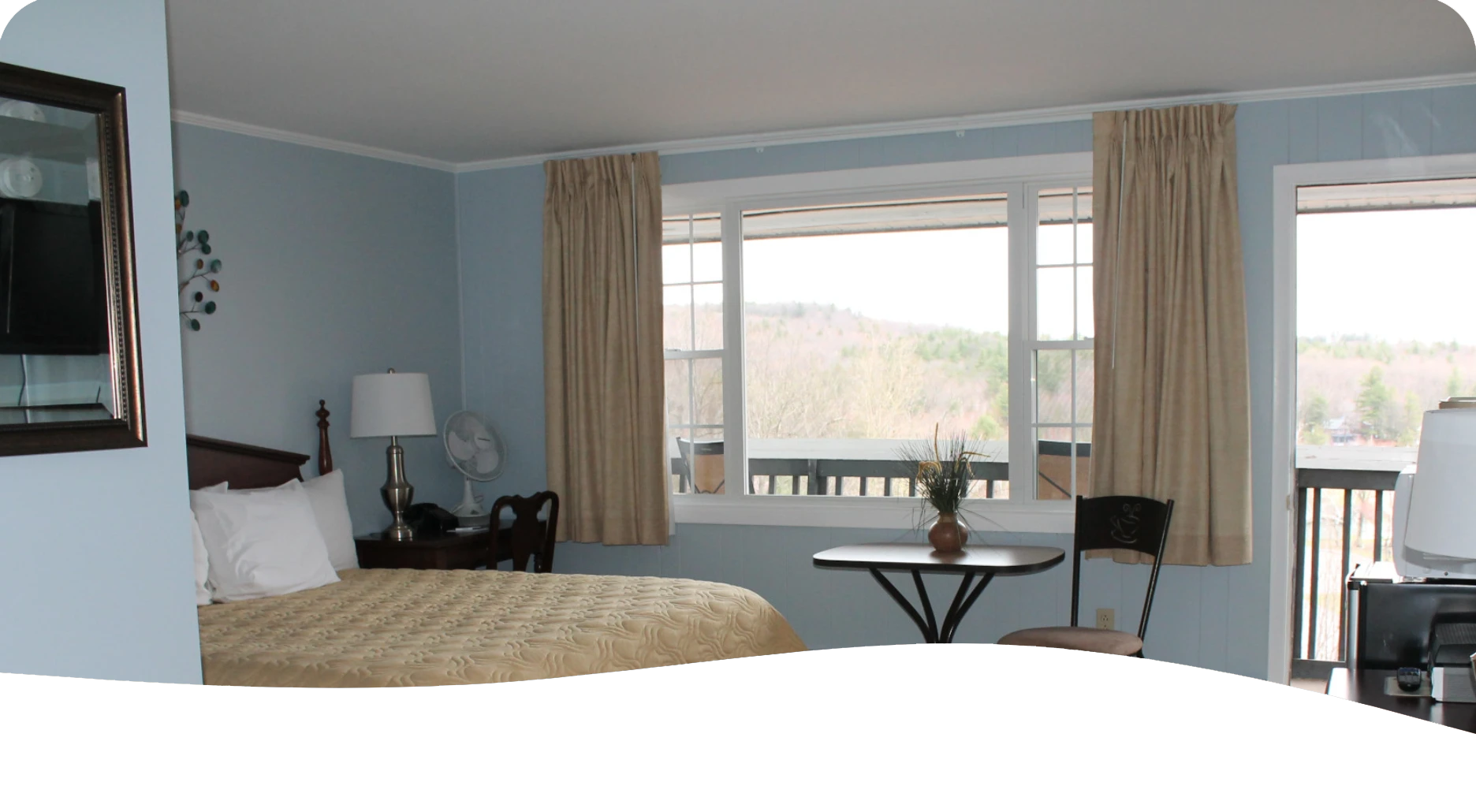 Bedroom with bed, desk, table, window overlooking trees, beige curtains.
