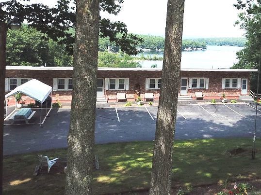 A brick motel with parking spaces in front, viewed through trees, with a lake visible in the background.