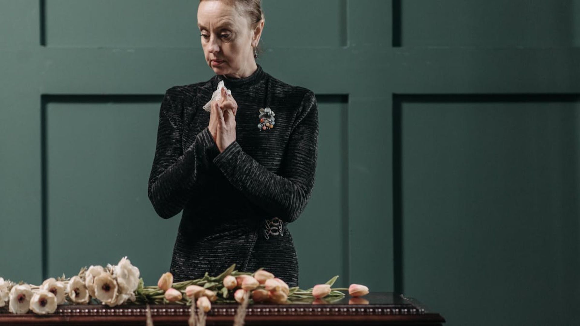 Una persona vestida de negro permanece de pie con las manos entrelazadas sobre unas flores colocadas sobre una mesa, apoyada contra una pared revestida de paneles verdes.