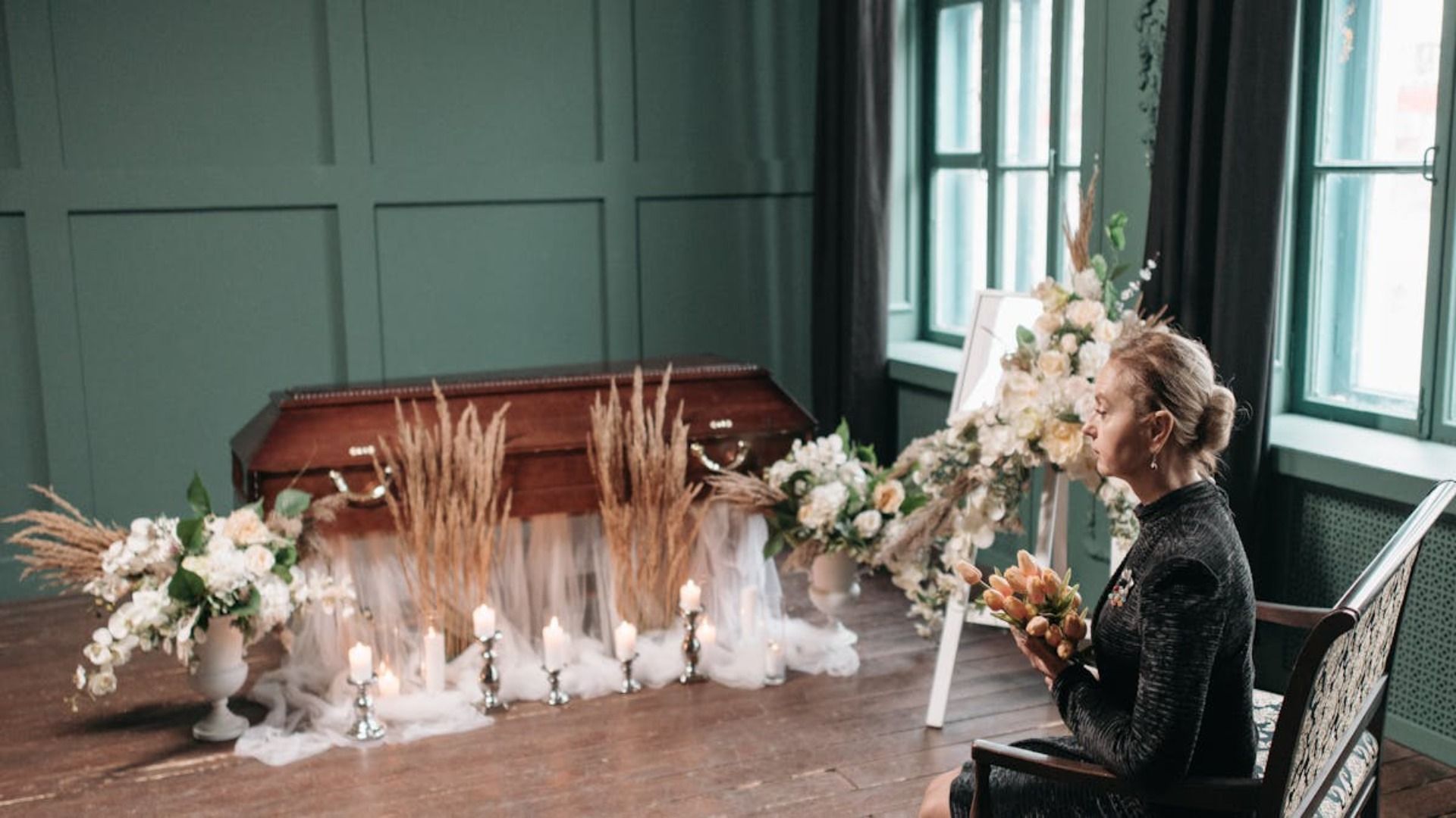 En una habitación, una persona vestida de traje formal negro está sentada junto a un ataúd de madera, rodeada de arreglos florales blancos y velas.