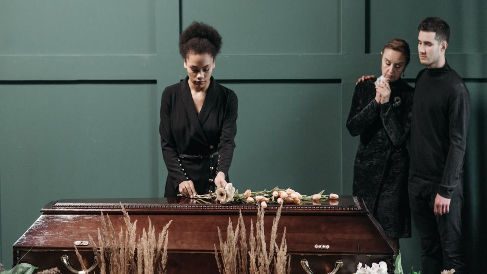 Tres personas vestidas de negro permanecen de pie en un ambiente sombrío alrededor de un ataúd de madera, una de ellas colocando flores sobre la tapa.